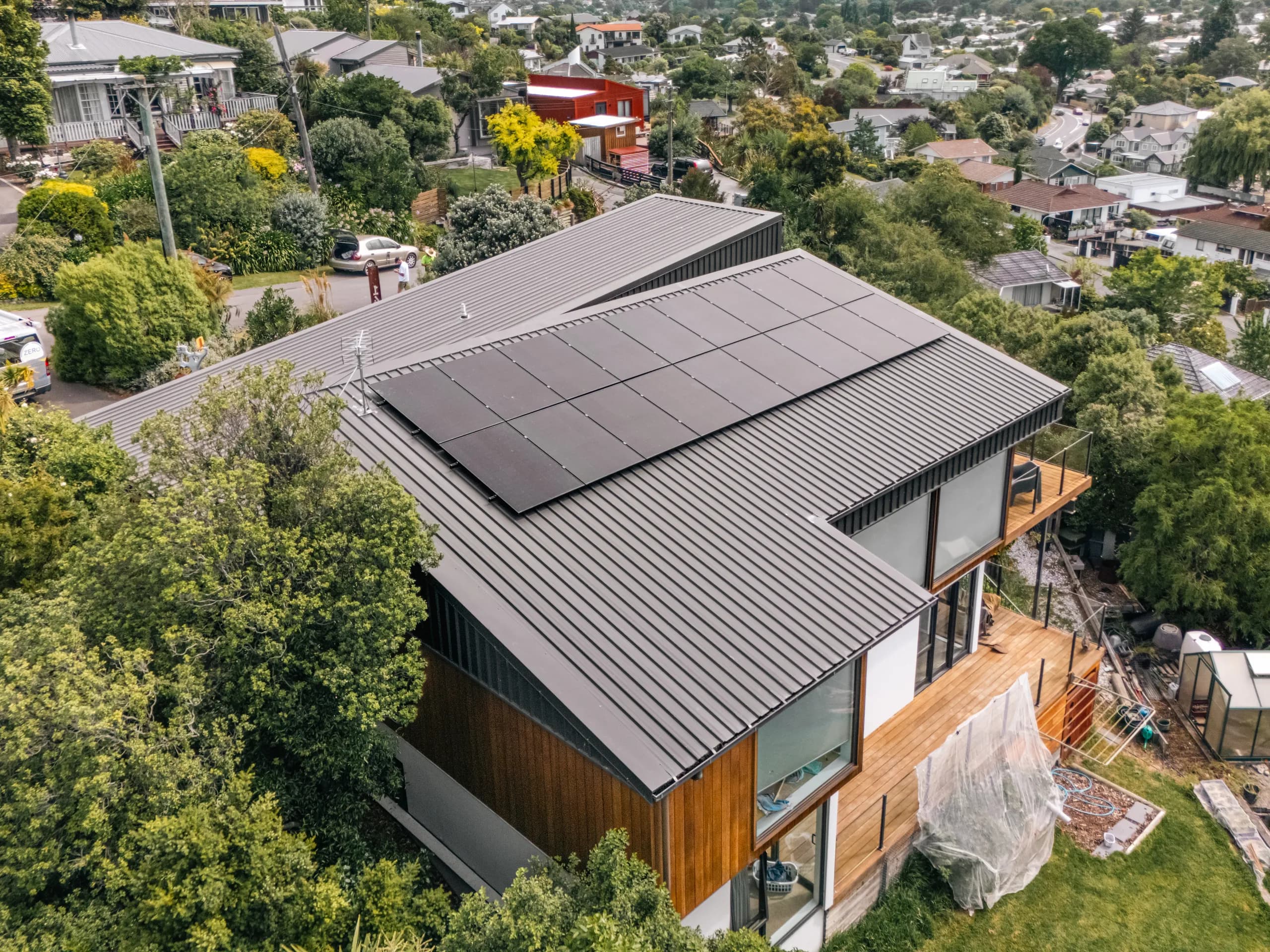 Drone shot of a modern two-storey timber-and-black-clad home with black solar panels flush on its dark standing-seam roof, a ZERO branded van parked on the street below, in a leafy Christchurch suburb.