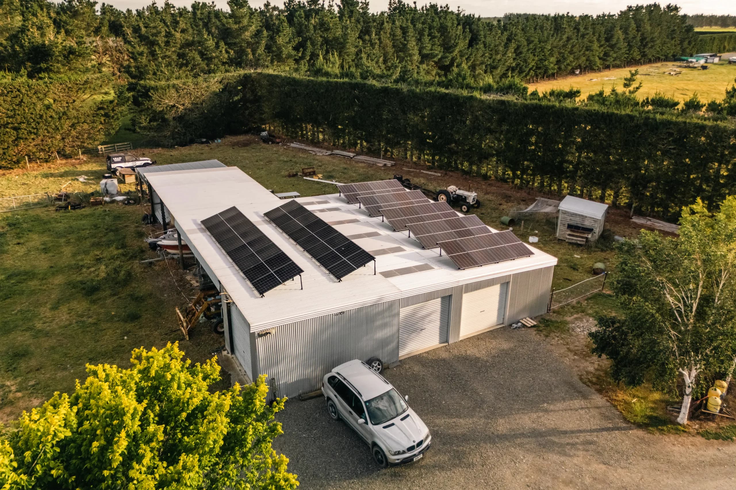 Golden-hour drone view of solar panels mounted on a white shed roof with a silver BMW SUV parked on the gravel below, bordered by tall hedging and pine shelterbelts.